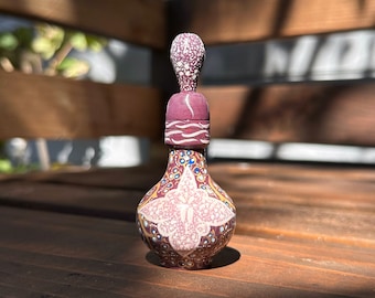 Puede incluir: Una pequeña botella de cerámica pintada a mano con un tapón decorativo. La botella está decorada con un diseño floral rosa y blanco, y el tapón está decorado con puntos blancos. La botella está sentada sobre una superficie de madera.