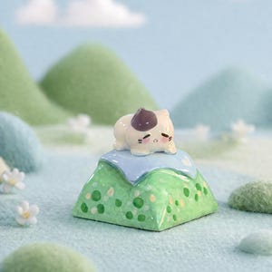 May include: A whimsical ceramic figurine of a sleeping cat with a brown spot on its head, resting on a green mountain with white and yellow accents. The mountain sits on a blue base, with a soft, pastel-colored background.