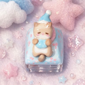 May include: A light blue nail polish bottle with a decorative cat figurine wearing a blue hat and bib. The cat is light brown with pink accents. The background includes pink stars and clouds.