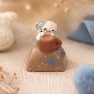 May include: A small, glossy, tan-colored artisan keycap featuring a sculpted white and gray cat figure holding a brown coffee mug. The base of the keycap is decorated with small, light blue four-pointed star shapes and tiny white dots.
