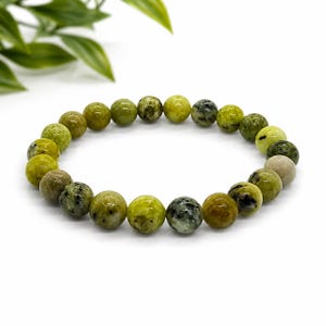May include: A stretch bracelet composed of round, polished beads in varying shades of lime green, olive, and chartreuse. The beads feature natural mottled patterns, dark speckles, and translucent inclusions, arranged in a circular loop.