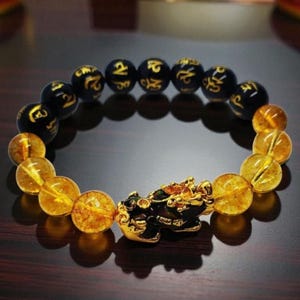May include: A beaded bracelet featuring a mix of round black beads with gold-colored inscriptions and translucent yellow beads. The bracelet includes a central gold-colored charm shaped like a mythical creature, positioned between the two bead types.