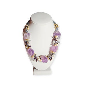 May include: A statement necklace featuring large, raw, light purple amethyst stones interspersed with smaller dark blue beads, white pearls, and gold-toned metal accents. The design includes clusters of mixed beads and stones arranged symmetrically.