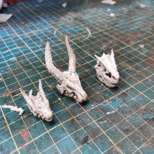 Puede incluir: Tres miniaturas de calaveras de dragón blancas con estructuras óseas detalladas están dispuestas sobre una superficie de cuadrícula azul y verde. Las calaveras varían en tamaño, una de ellas con grandes cuernos. La escena sugiere un tema de fantasía o artesanía.