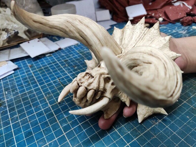 Puede incluir: Un cr&aacute;neo blanco esculpido detallado con dos cuernos grandes. El cr&aacute;neo tiene una superficie texturizada y parece estar hecho de resina.