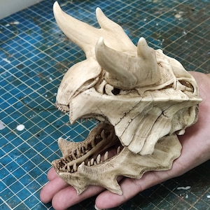 Può includere: Un teschio di drago scolpito, dettagliato, di colore avorio, con la mascella aperta, che rivela file di denti. Il teschio ha due grandi corna sulla parte superiore e dettagli intricati in tutto.