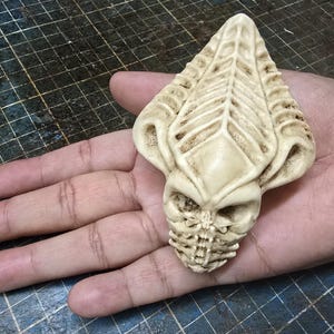 May include: A cream-colored, intricately carved sculpture of a skull-like alien head. The detailed design features a prominent forehead crest, eye sockets, and a segmented jaw. The sculpture is held in a hand.