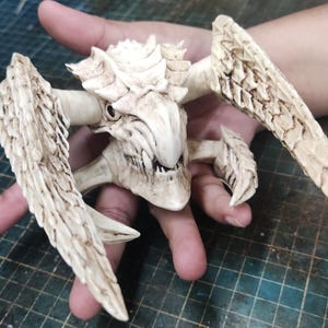 Può includere: Una scultura dettagliata, bianco sporco, di una creatura con testa cornuta, grandi ali e molteplici appendici. La scultura è tenuta in una mano, mostrando il suo design e la sua consistenza intricati. Il volto della creatura ha denti affilati.