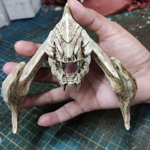 Può includere: Una testa di creatura scheletrica bianca dettagliata con una grande bocca aperta e denti affilati. La testa è scolpita con dettagli intricati e sembra essere fatta di resina.