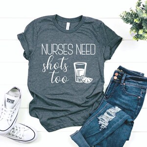 Puede incluir: Una camiseta gris oscuro con el texto "NURSES NEED shots too" en letras blancas. Un contorno blanco de un vaso de chupito con una rodaja de lima está al lado del texto.