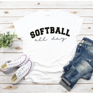 Op de afbeelding: Wit T-shirt met zwarte tekst die "Softball all day" luidt op een witte houten achtergrond. Het shirt is gestyled met een blauwe jeans en witte sneakers.