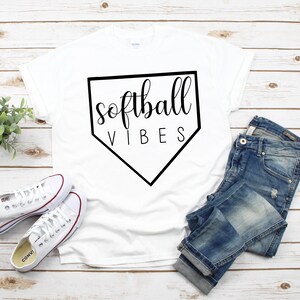 May include: White t-shirt with a black baseball diamond graphic and the text "softball vibes" printed on it. The shirt is laid out on a wooden surface with a pair of white sneakers and blue jeans.
