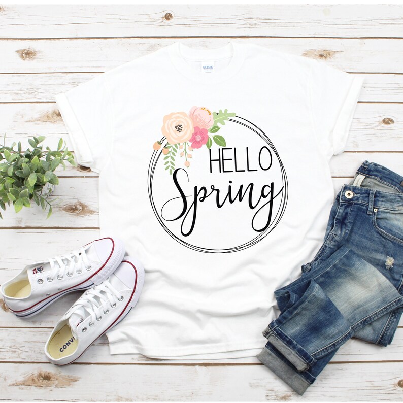 May include: White t-shirt with a floral wreath and the text "Hello Spring" printed on the front. The shirt is laid out on a wooden surface with a pair of white sneakers and blue jeans.