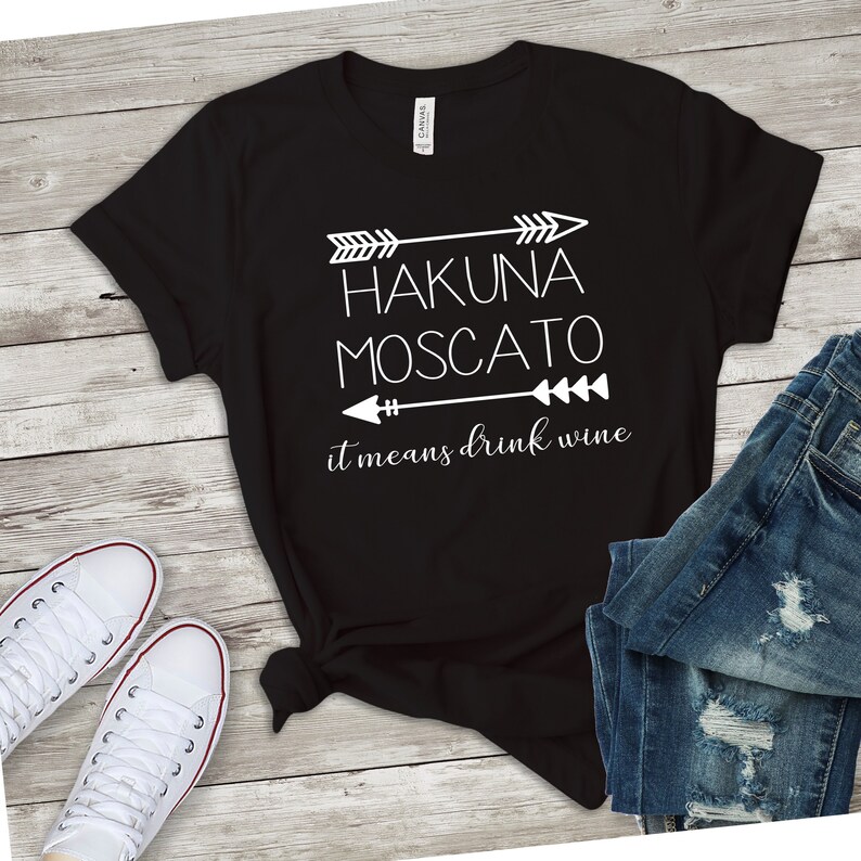 May include: Black t-shirt with white text that reads "Hakuna Moscato it means drink wine" with arrows on either side of the text.
