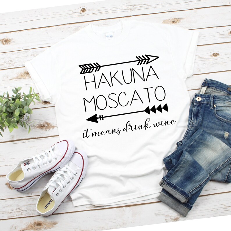 May include: White t-shirt with black text that reads "Hakuna Moscato it means drink wine" with arrows on either side of the text.