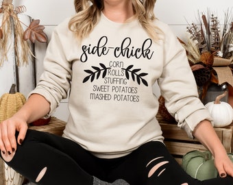 May include: A beige sweatshirt with the text "side-chick" in black cursive lettering. Below the text is a wreath of leaves with the words "corn rolls stuffing sweet potatoes mashed potatoes" written inside.