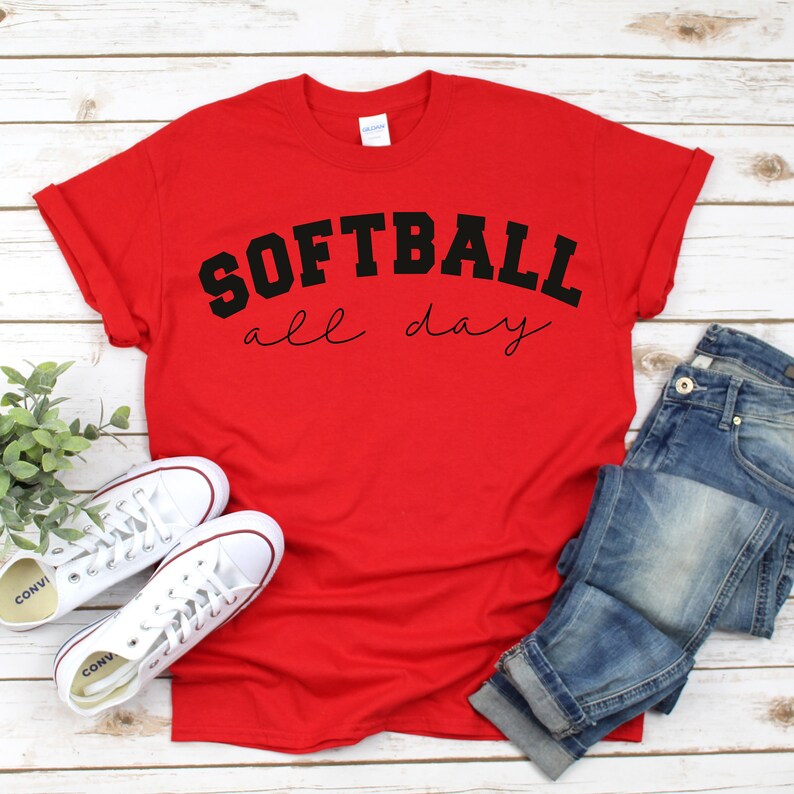Op de afbeelding: Rood T-shirt met zwarte tekst "Softball all day" op een witte houten achtergrond.