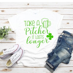 Puede incluir: Camiseta blanca con texto verde que dice "Take a Pitcher It Lasts Longer" y una ilustración verde de una jarra de cerveza con un trébol.