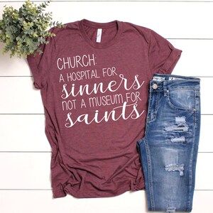May include: A maroon t-shirt with white text that reads "CHURCH: A HOSPITAL FOR SINNERS NOT A MUSEUM FOR SAINTS". The shirt is paired with blue jeans.