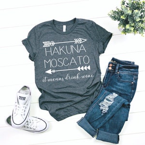 May include: Gray t-shirt with white text that reads "Hakuna Moscato it means drink wine" with arrows on either side of the text.
