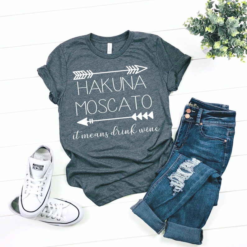 May include: Gray t-shirt with white text that reads "Hakuna Moscato it means drink wine" with arrows on either side of the text.