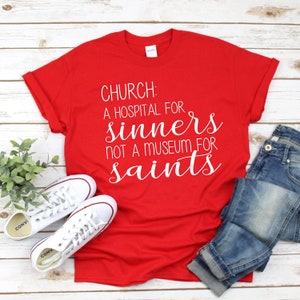 May include: Red t-shirt with white text that reads "CHURCH: A HOSPITAL FOR SINNERS NOT A MUSEUM FOR SAINTS". The shirt is laid flat on a wooden surface with a pair of white sneakers and blue jeans.