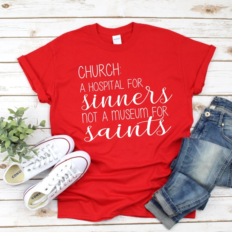 May include: Red t-shirt with white text that reads "CHURCH: A HOSPITAL FOR SINNERS NOT A MUSEUM FOR SAINTS". The shirt is laid flat on a wooden surface with a pair of white sneakers and blue jeans.