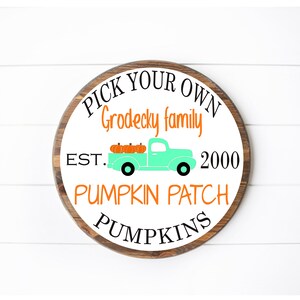 Puede incluir: Un letrero redondo de madera blanca con borde marrón que dice "Pick Your Own Grodecky Family Est. 2000 Pumpkin Patch Pumpkins". Una camioneta azul verdosa con calabazas naranjas en la caja está en el centro del letrero.