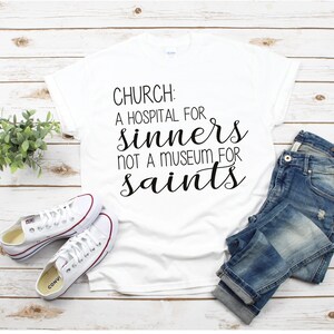 May include: White t-shirt with black text that reads "CHURCH: A HOSPITAL FOR SINNERS NOT A MUSEUM FOR SAINTS". The shirt is laid flat on a wooden surface with a pair of white sneakers and blue jeans.