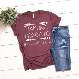 May include: A maroon t-shirt with white text that reads "Hakuna Moscato it means drink wine" with arrows on either side of the text.
