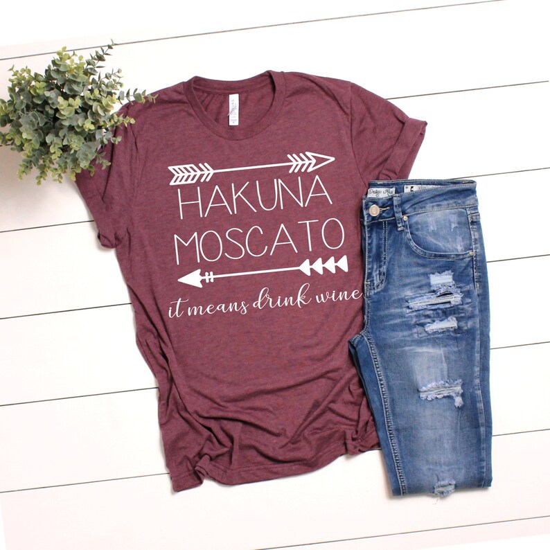 May include: A maroon t-shirt with white text that reads "Hakuna Moscato it means drink wine" with arrows on either side of the text.