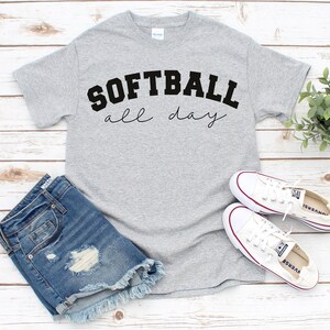 Op de afbeelding: Grijs T-shirt met de tekst "SOFTBALL all day" in zwarte letters. Het shirt wordt gecombineerd met een spijkerbroekje en witte sneakers.