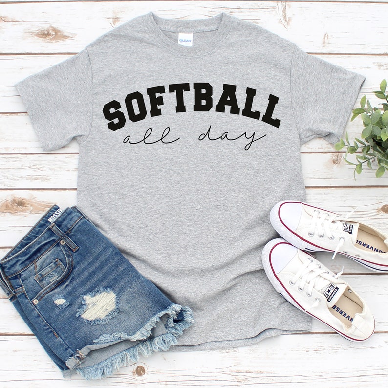 Op de afbeelding: Grijs T-shirt met de tekst "SOFTBALL all day" in zwarte letters. Het shirt wordt gecombineerd met een spijkerbroekje en witte sneakers.