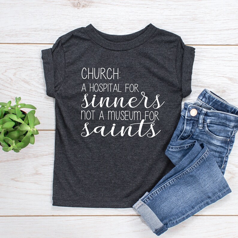 May include: A dark gray t-shirt with white text that reads "CHURCH: A HOSPITAL FOR SINNERS NOT A MUSEUM FOR SAINTS". The shirt is laying on a white wooden surface with a pair of blue jeans folded next to it.