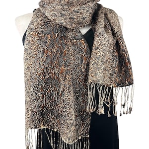 May include: A brown and tan patterned scarf with a snake skin design is draped over a black dress form. The scarf has a fringed edge with small tassels. The scarf is made of a soft material.