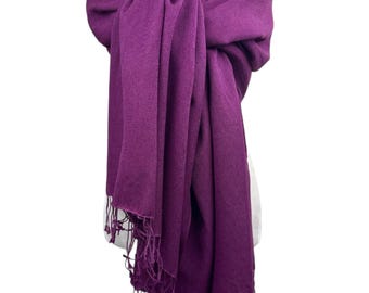 Pode incluir: Um cachecol roxo escuro com uma borda com franjas é exibido em um manequim. O cachecol é enrolado ao redor do pescoço e dos ombros, com as pontas penduradas. Um colar de pérolas é visível no pescoço.