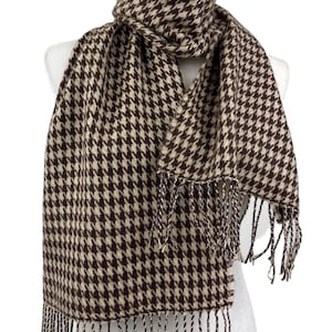 May include: A rectangular scarf featuring a brown and beige houndstooth pattern. The scarf is draped around a mannequin neck and includes twisted fringe detailing along the bottom edges, creating a textured finish to the woven fabric design.