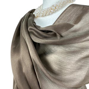 Pode incluir: Um len&ccedil;o cor de taupe com um brilho subtil drapeado sobre um manequim branco. O len&ccedil;o apresenta uma textura suave e fluida, combinado com um colar de v&aacute;rias voltas composto por numerosas pequenas p&eacute;rolas brancas redondas em design torcido.
