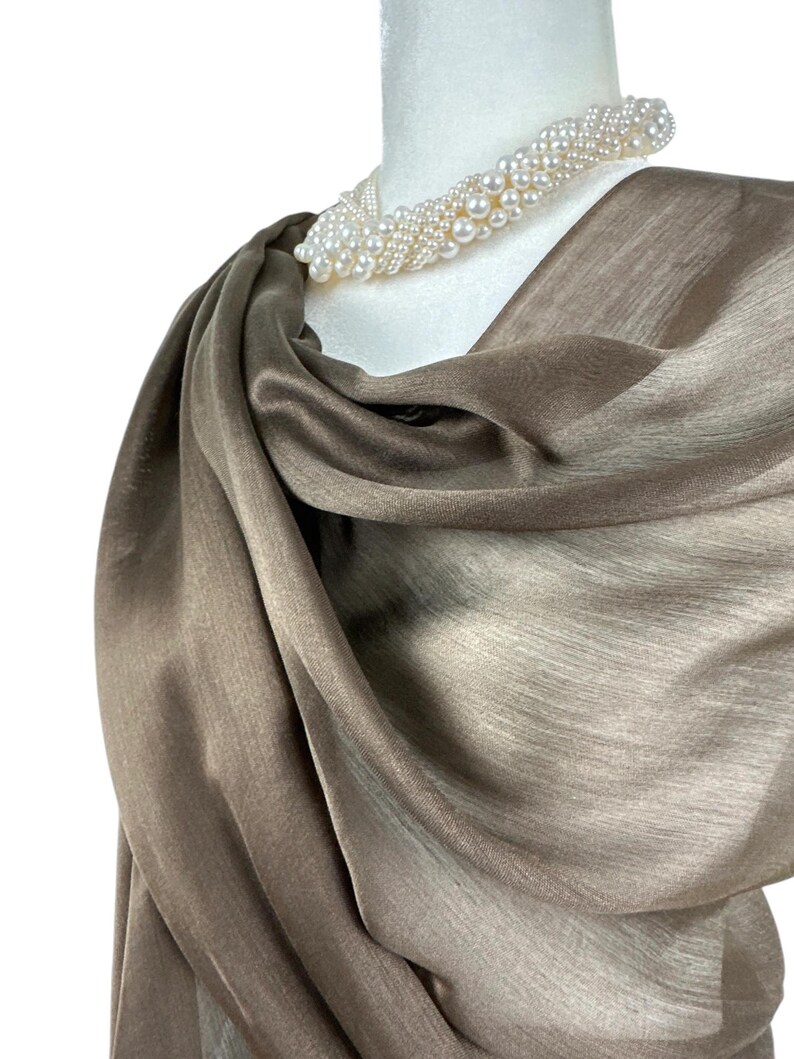 Pode incluir: Um len&ccedil;o cor de taupe com um brilho subtil drapeado sobre um manequim branco. O len&ccedil;o apresenta uma textura suave e fluida, combinado com um colar de v&aacute;rias voltas composto por numerosas pequenas p&eacute;rolas brancas redondas em design torcido.
