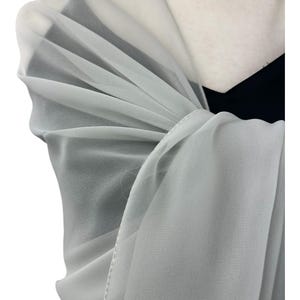 May include: A light gray, semi-sheer chiffon shawl draped over the shoulders. The fabric is lightweight and translucent, featuring a delicate, fine-stitched hem along the edge. The shawl is layered over a solid black garment, creating a soft contrast.
