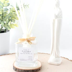 May include: A clear glass diffuser bottle with reeds, labeled "Fields of Fatima", sits on a wooden coaster. A white ceramic statue and a potted plant with green and yellow leaves are also in the image. The bottle is tied with a beige ribbon.