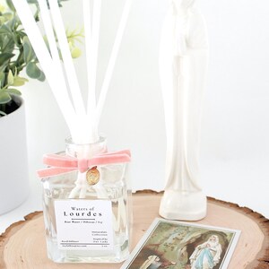 May include: A clear glass reed diffuser with a pink ribbon and gold charm, labeled "Waters of Lourdes." A white ceramic statue and a religious card are on a wooden slice. Decorative white reeds are in the diffuser.