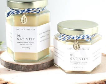 May include: Two hexagonal glass jars filled with light yellow wax, each featuring a gold metal lid, a blue and white twine tie, and a small gold religious charm. The white labels read Joyful Mysteries 03. Nativity, Frankincense, Myrrh, Amber, Incense.