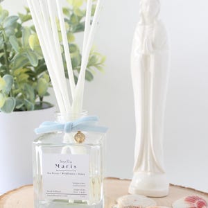 May include: A clear glass reed diffuser with white reeds, a light blue ribbon, and a gold charm. The label reads "Stella Maris." A white ceramic statue and seashells rest on a wooden slice. A potted plant is in the background.
