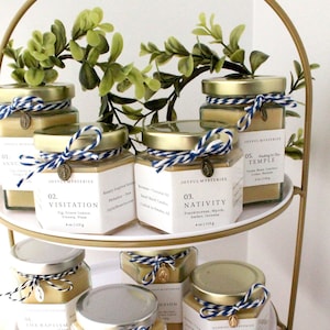 Puede incluir: Un soporte de metal dorado de dos niveles muestra una colección de pequeños frascos de vidrio con tapas doradas. Cada frasco contiene una vela y está decorado con una cinta azul y blanca y un pequeño amuleto. Los frascos están etiquetados con números y títulos relacionados con la fe católica, como "Anunciación", "Visitación", "Natividad", "El Bautismo", "Bodas de Caná", "Asunción", "Su Pasión", y "Encontrado en el Templo".