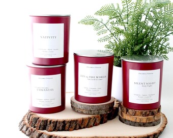 May include: Four red Christmas candles with white labels and silver lids are displayed on a wooden slab and coasters. The labels read "Nativity", "O Come, Emmanuel", "Joy to the World", and "Silent Night". A green plant is in the background.