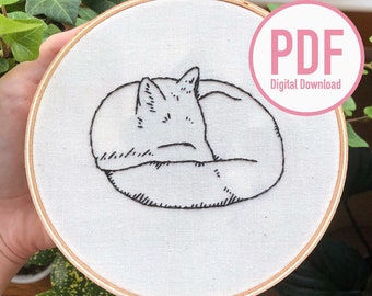 Könnte beinhalten: Eine Schwarzweiß-Zeichnung eines zusammengerollten schlafenden Katers, gestickt auf einem weißen Stoff in einem hölzernen Stickrahmen. Der Rahmen wird von einer Hand gehalten. Das Bild enthält auch den Text "PDF Digitaler Download".
