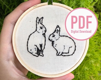 Könnte beinhalten: Nahaufnahme einer Hand, die einen kleinen Stickrahmen mit zwei schwarzen gestickten Kaninchen auf einem weißen Stoffhintergrund hält. Der Rahmen ist aus Holz und hat einen Metallständer. Der Text "PDF Digitaler Download" befindet sich auf einem rosa Kreis in der oberen rechten Ecke des Bildes.