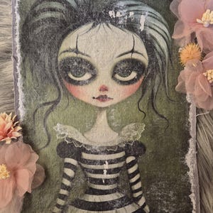 May include: A rectangular fabric art piece featuring a stylized illustration of a doll-like figure with large eyes and dark makeup. The figure wears a black and white striped dress with a ruffled collar. The artwork has a distressed, vintage aesthetic.