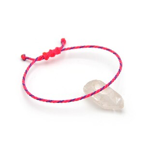Pode incluir: Uma pulseira ajustável feita à mão com um cordão trançado rosa e roxo vibrante. A pulseira é amarrada nas extremidades para ajuste. Um cristal de quartzo transparente está ao lado da pulseira em um fundo branco. Joias.